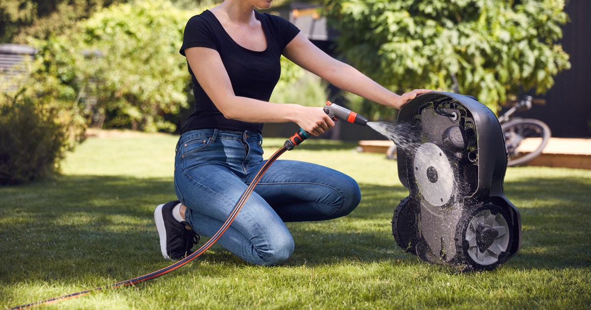 Fotografía oficial de una persona lavando con una manguera la parte inferior de un Husqvarna Automower, sostenido verticalmente sobre el césped.