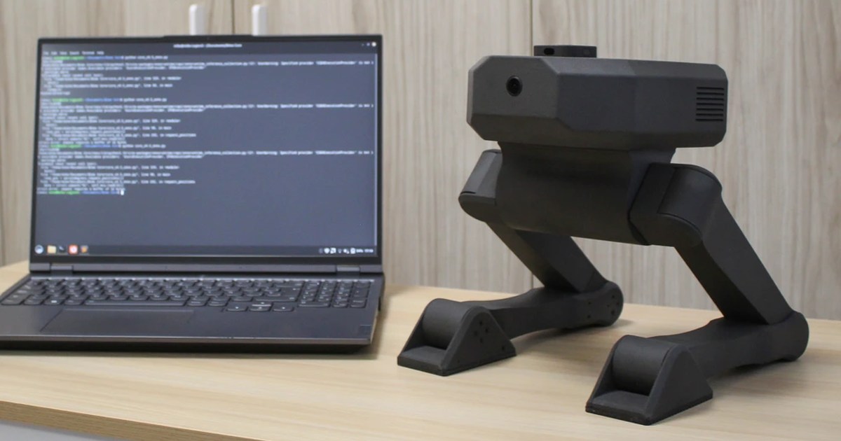 Fotografía oficial del robot bípedo Bimo sentado sobre una mesa junto a un ordenador portátil.