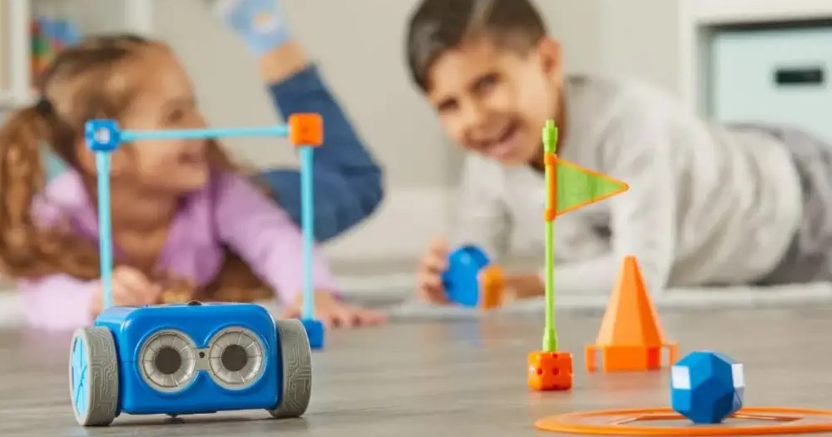 Botley 2.0 azul en primer plano sobre un suelo de madera, con un circuito de piezas y dos niños jugando al fondo; fotografía de producto distribuida por RobotShop.