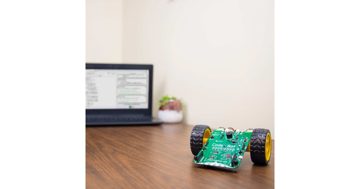 CodeBot con placa electrónica verde y dos ruedas sobre una mesa junto a un ordenador.