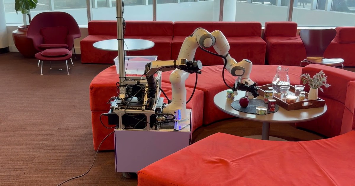 Frame oficial de COWBOY: un robot con base de ruedas y brazo blanco acerca su mano a una mesa con objetos, rodeada de sofás rojos.