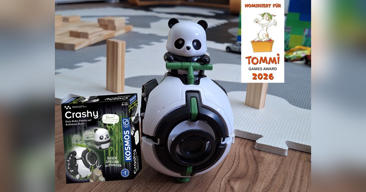 Fotografía promocional oficial de Crashy completo sobre el suelo, junto a su caja y el anuncio de su nominación al TOMMI 2026.
