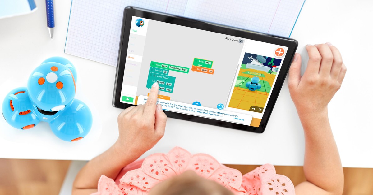Fotografía oficial de un robot Dash azul junto a una niña que usa programación por bloques en una tableta; la pantalla muestra también un Dash virtual.