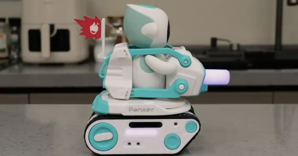 Fotograma oficial de un robot Eilik montado en un Panxer blanco y turquesa, con orugas y una bandera roja, sobre una superficie de cocina.