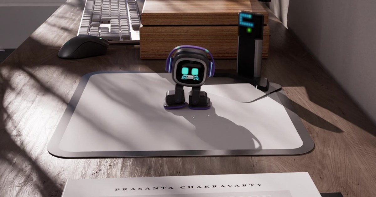Frame oficial de EMO de pie sobre una alfombrilla blanca en un escritorio de madera, con Home Station y un teclado detrás.