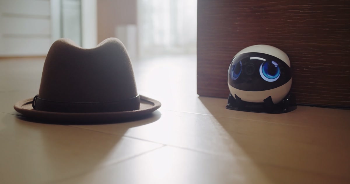 EBO Air 2 Plus blanco en su base de carga junto a un sombrero sobre el suelo, fotograma del vídeo promocional oficial de Enabot.