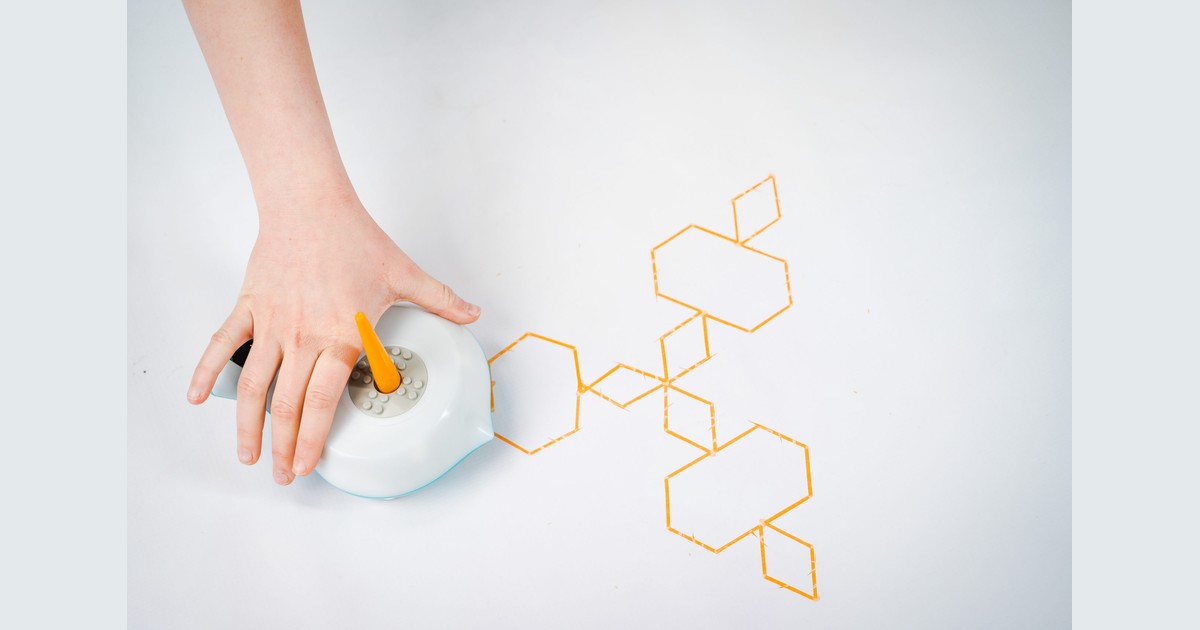Una mano sobre un Finch 2.0 con un rotulador naranja en el soporte, junto a figuras geométricas dibujadas sobre papel blanco; fotografía oficial.