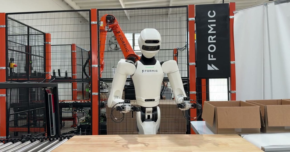 Imagen oficial de Formic y Physical Intelligence: un humanoide blanco ante una mesa y cajas abiertas, con un brazo industrial naranja tras una valla.