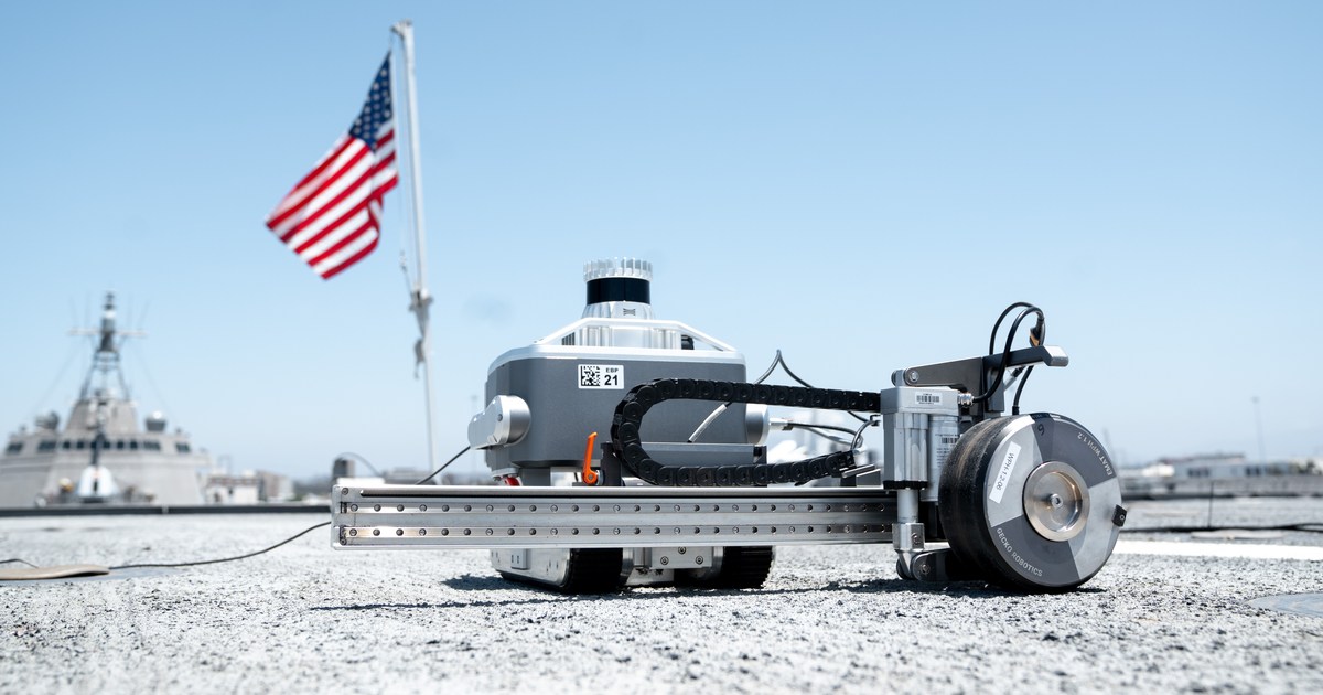Fotografía de Gecko Robotics del robot Komodo sobre una cubierta naval, con su mecanismo de inspección extendido y una bandera estadounidense al fondo.