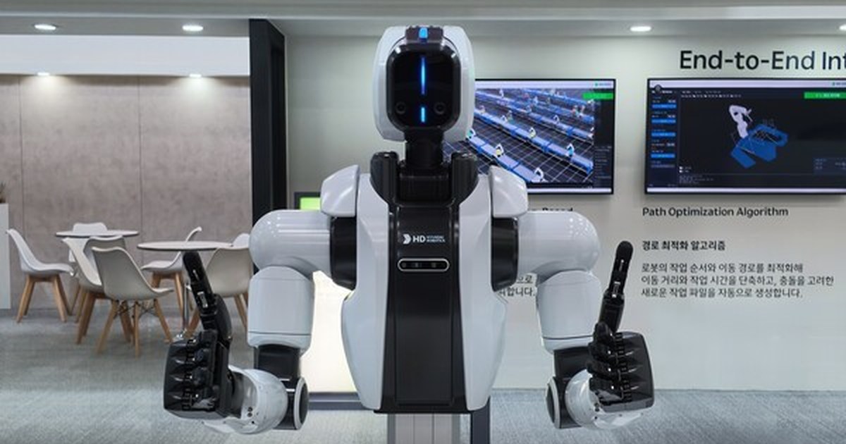 Fotografía oficial de HD Hyundai: su robot blanco de dos brazos, instalado sobre un soporte, levanta ambos pulgares en el stand de ROBEX 2026.