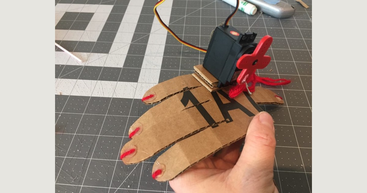 Una mano de cartón con hilos rojos y un servomotor sujeto al dorso, sostenida sobre una base de corte; fotografía del tutorial oficial de BirdBrain.