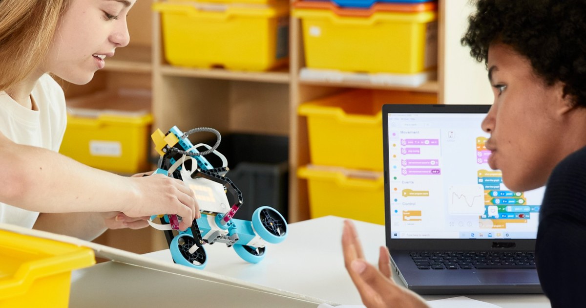 Dos estudiantes trabajan con un robot LEGO Education SPIKE Prime de ruedas y un portátil con bloques de programación, en una fotografía oficial.