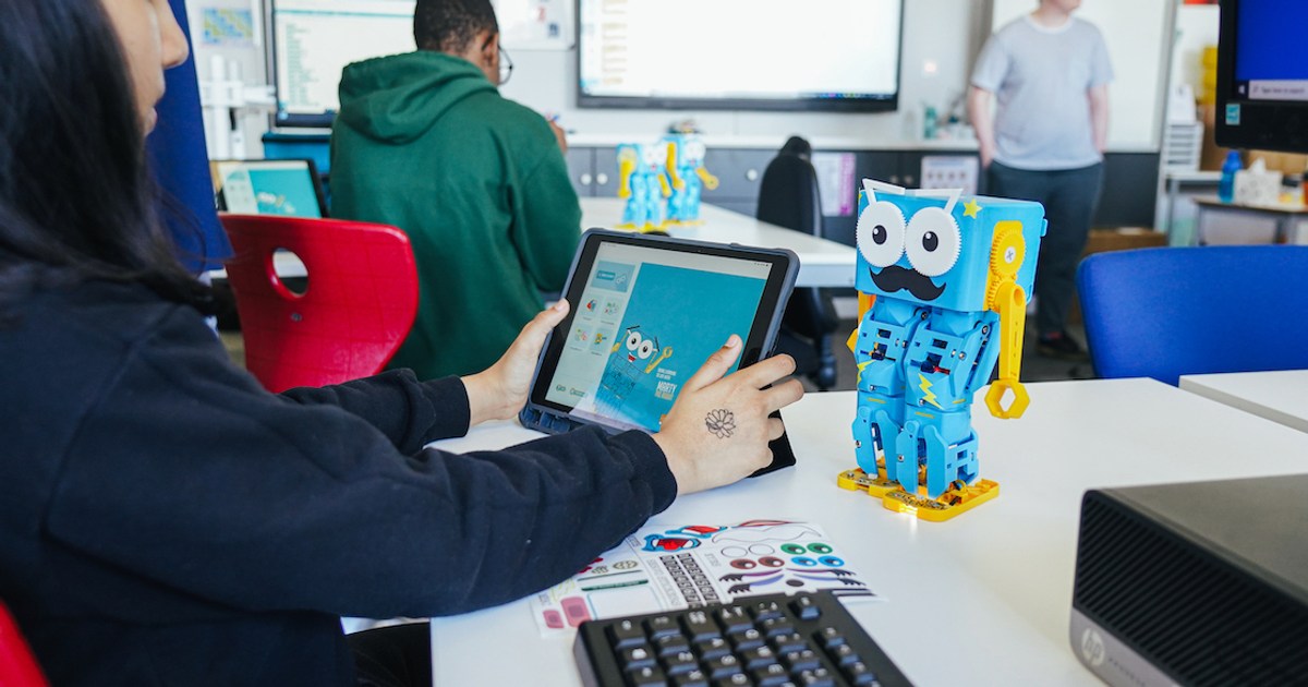 Fotografía oficial de Marty V2 azul y amarillo sobre una mesa de aula, junto a una persona que utiliza su interfaz de programación en una tableta.
