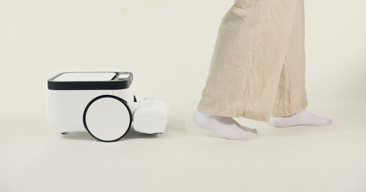 Fotografía promocional oficial del robot Matic blanco, visto de lado junto a los pies de una persona con pantalones claros y calcetines.