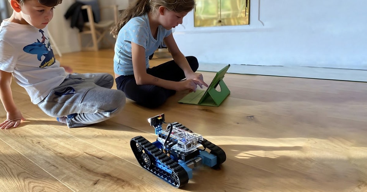 Fotografía oficial de un mBot Ranger azul con orugas sobre un suelo de madera, junto a dos niños y una tableta.