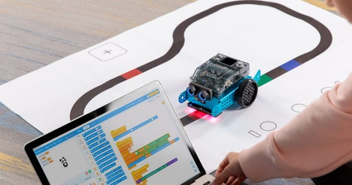 Robot Makeblock mBot2 sobre una pista de líneas junto a un portátil con programación por bloques, en una fotografía oficial de una actividad.