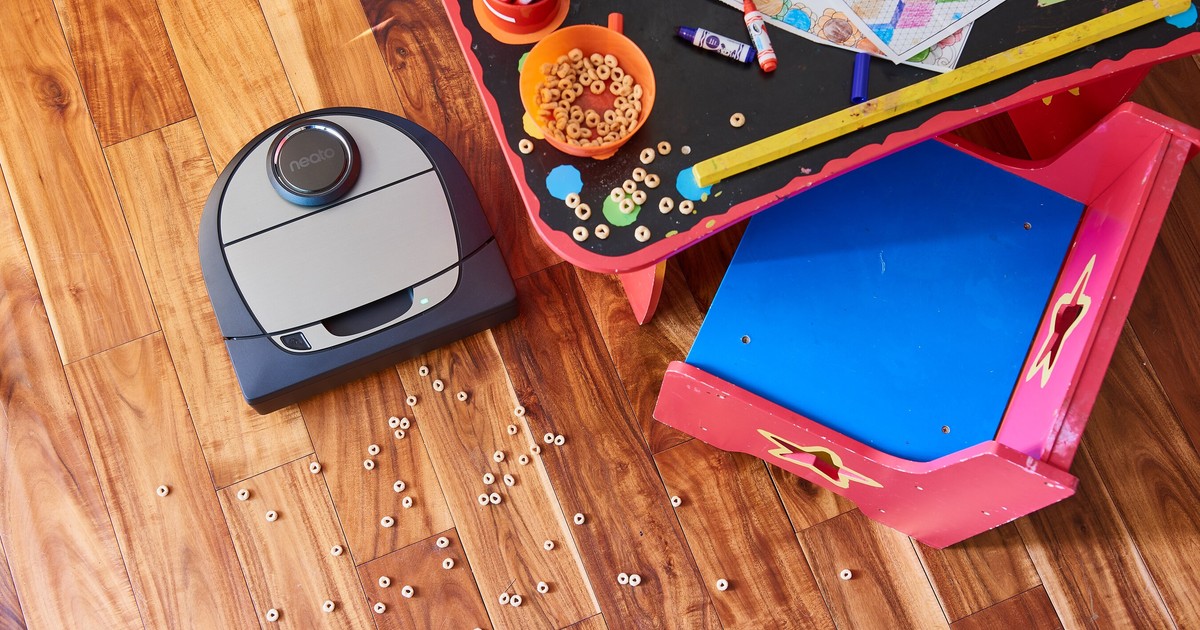 Fotografía oficial de un Neato Botvac D7 Connected sobre un suelo de madera, junto a cereales derramados y una mesa infantil.