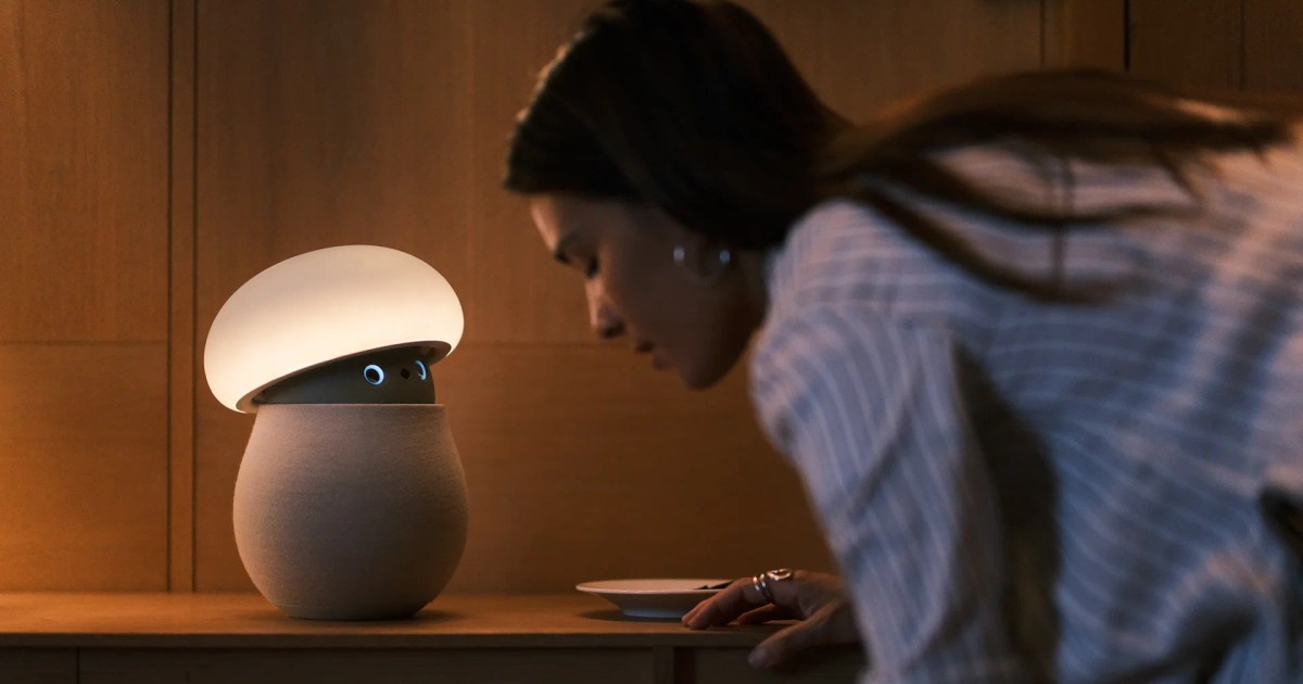 Fotografía oficial de Mori iluminado sobre una mesa, con la cabeza inclinada y los ojos azules visibles, mientras una mujer se acerca al robot.