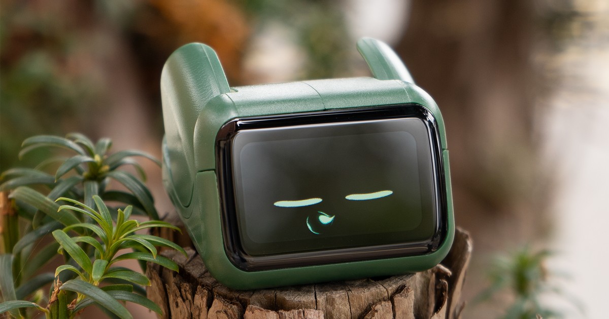 Fotografía oficial de un Ortomi Umi verde sobre un tronco, junto a plantas, con ojos y una boca dibujados en su pantalla y las dos orejas visibles.