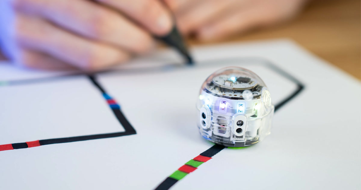 Fotografía oficial de un Ozobot Evo transparente sobre una línea con códigos de colores, mientras una mano dibuja el recorrido al fondo.