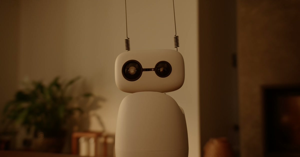 Reachy Mini blanco sobre una mesa, con la cabeza levantada y plantas al fondo, en un fotograma del vídeo oficial de Pollen Robotics.