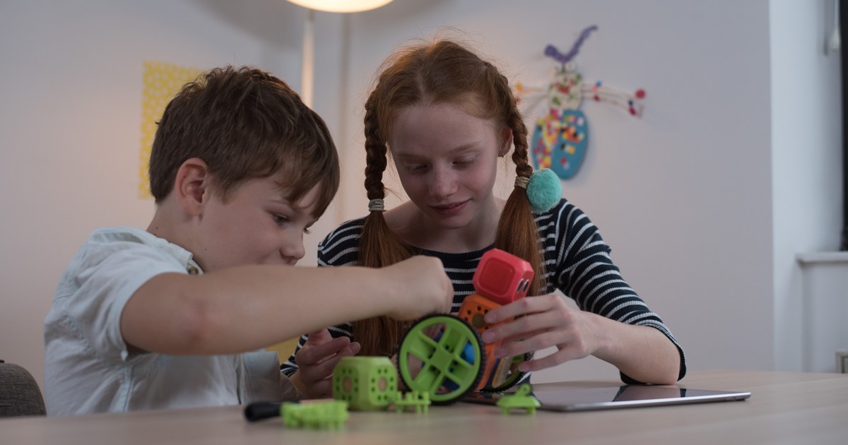 Fotografía oficial de dos niños montando un robot Robo Wunderkind con bloques de colores y ruedas verdes, junto a una tableta sobre la mesa.
