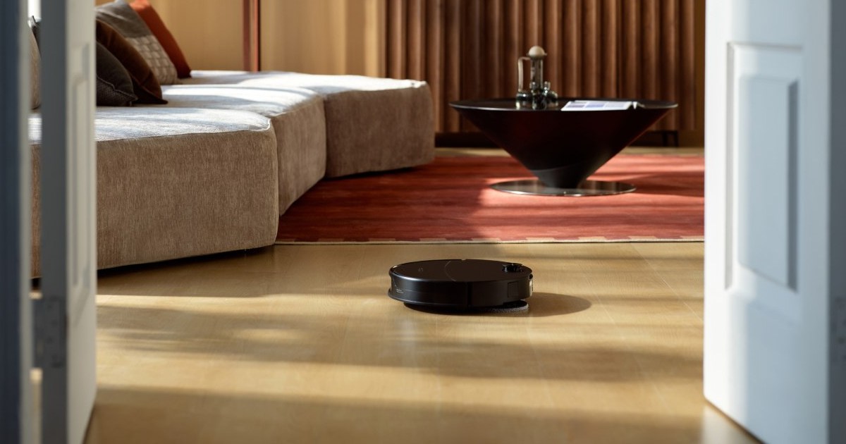 Fotografía promocional oficial de un Roborock Saros 20 Neo negro sobre suelo de madera, visto a través de una puerta frente a un sofá y una alfombra.