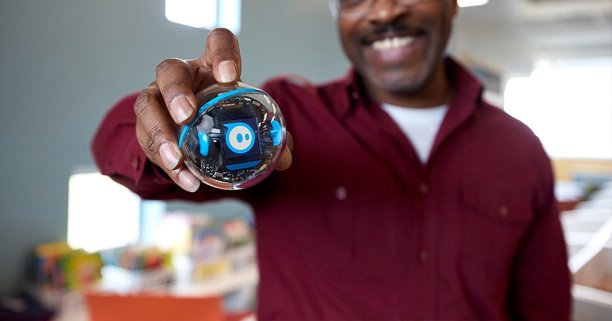 Fotografía oficial de una persona sosteniendo un Sphero BOLT+ transparente, con una cara azul en su pantalla, en un aula.