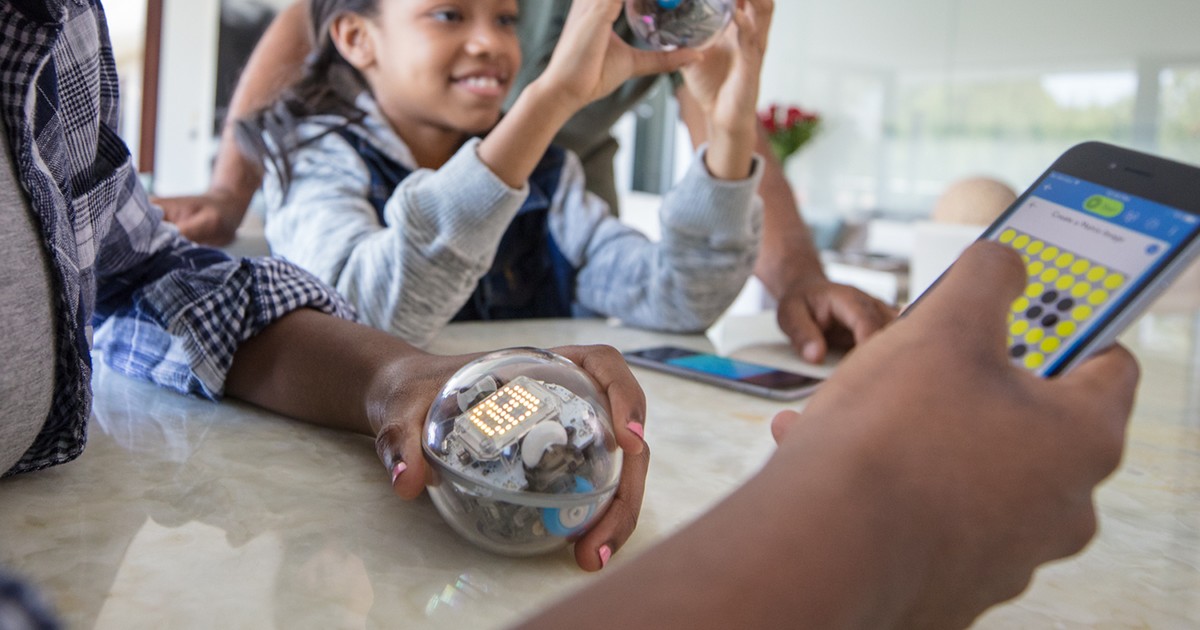 Fotografía oficial de un Sphero BOLT sostenido sobre una mesa junto a una tableta que muestra un editor de su matriz LED.