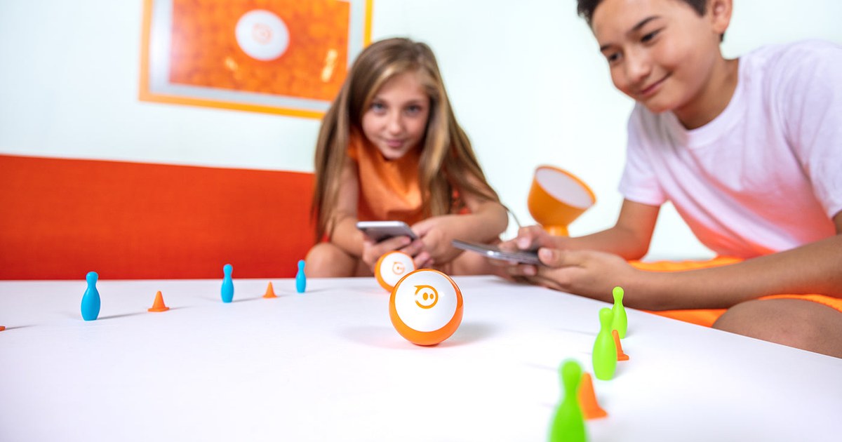 Dos niños controlan robots Sphero Mini naranjas con dispositivos móviles entre bolos y conos sobre una mesa, en una fotografía oficial de Sphero.