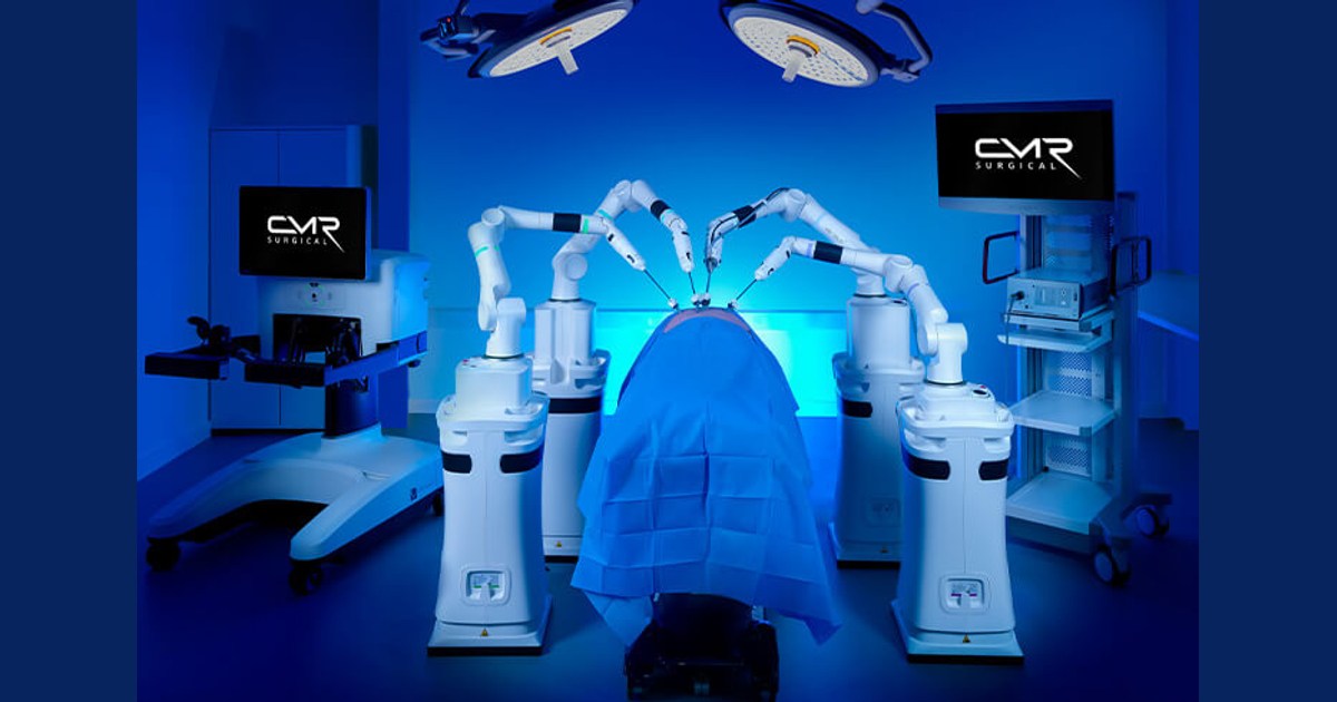 Fotografía oficial de Versius Plus en una sala iluminada en azul, con sus brazos independientes alrededor de una mesa cubierta y pantallas a ambos lados.
