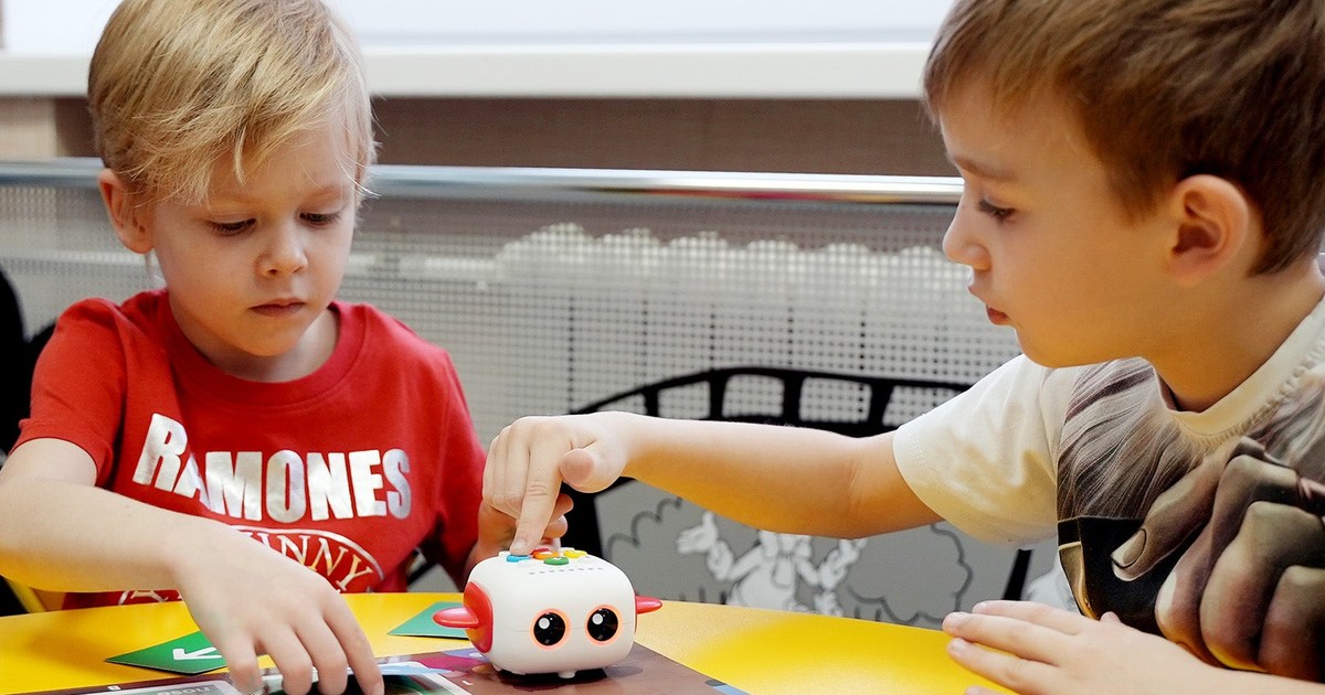 Dos niños programan un Tale-Bot Pro blanco con accesorios rojos sobre un tapete, en una fotografía oficial de MatataStudio.