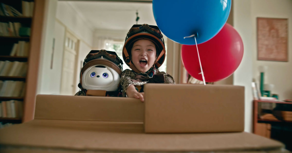 Imagen promocional oficial de TCL AiMe y un niño con cascos de aviador, dentro de una caja de cartón junto a globos.