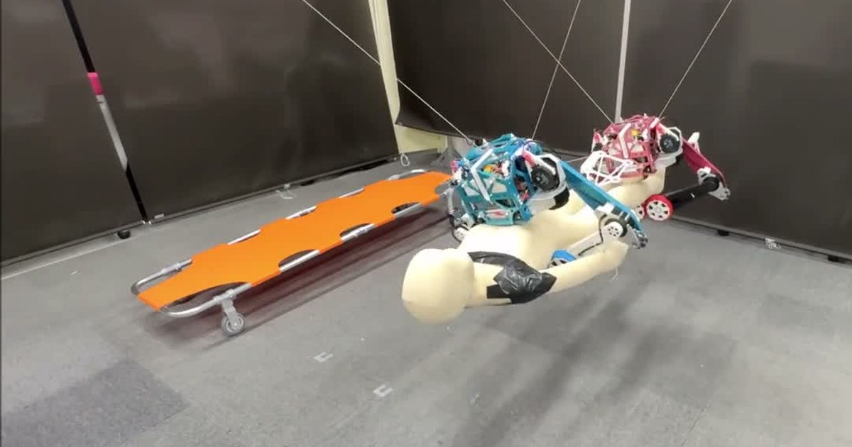 Fotograma oficial de TwinX: dos robots azul y rojo, suspendidos de cables, sostienen un maniquí junto a una camilla naranja en el laboratorio.