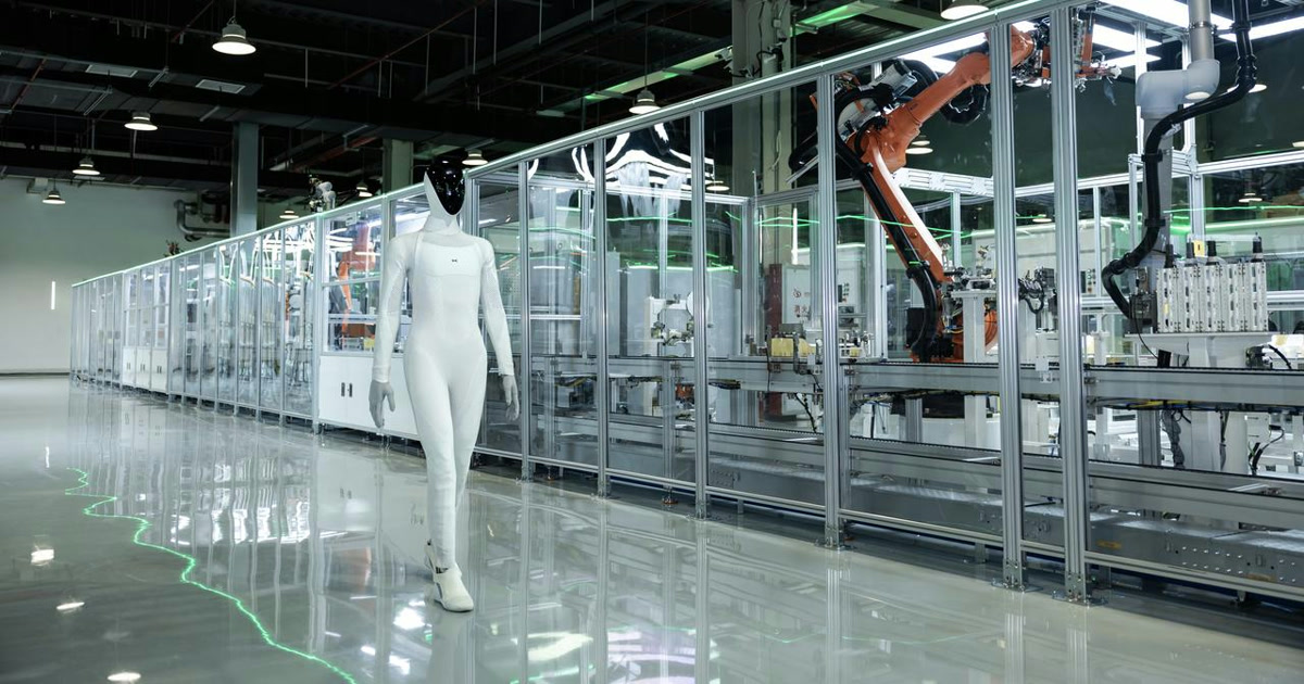 Fotografía oficial del humanoide XPENG IRON caminando junto a una línea de montaje con brazos industriales tras un cerramiento transparente.