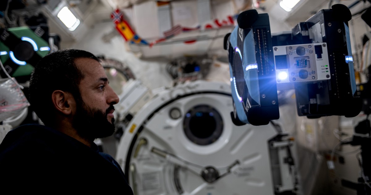 El astronauta Sultan Alneyadi observa un robot Astrobee azul flotando dentro de la Estación Espacial Internacional; otro robot verde aparece al fondo. Fotografía de la NASA de mayo de 2023.