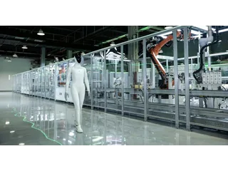 Fotografía oficial del humanoide XPENG IRON caminando junto a una línea de montaje con brazos industriales tras un cerramiento transparente.