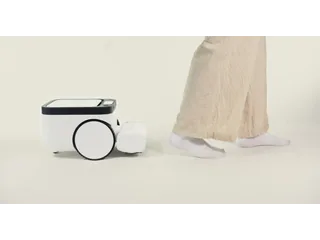 Fotografía promocional oficial del robot Matic blanco, visto de lado junto a los pies de una persona con pantalones claros y calcetines.