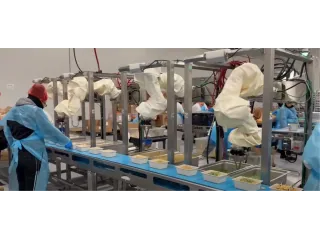 Robots de Chef Robotics con fundas protectoras trabajando junto a personas en una línea de preparación de bandejas de comida.