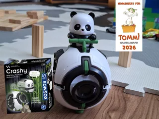 Fotografía promocional oficial de Crashy completo sobre el suelo, junto a su caja y el anuncio de su nominación al TOMMI 2026.