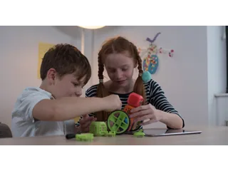 Fotografía oficial de dos niños montando un robot Robo Wunderkind con bloques de colores y ruedas verdes, junto a una tableta sobre la mesa.