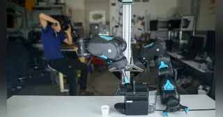 Blue en una demostración de teleoperación con sus dos brazos y un operador al fondo.
