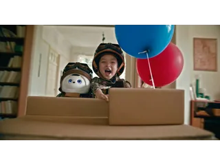 Imagen promocional oficial de TCL AiMe y un niño con cascos de aviador, dentro de una caja de cartón junto a globos.