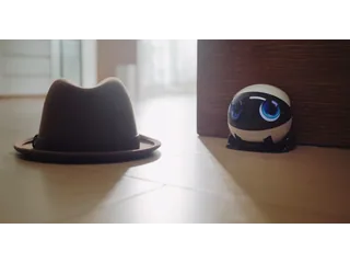 EBO Air 2 Plus blanco en su base de carga junto a un sombrero sobre el suelo, fotograma del vídeo promocional oficial de Enabot.
