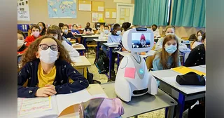 Buddy blanco con pantalla de videollamada junto a alumnos en un aula.