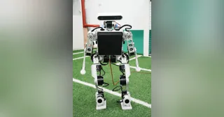 Fotografía oficial de MOS9.2 completo, de frente, con estructura metálica, brazos y pies sobre un campo de fútbol robótico.
