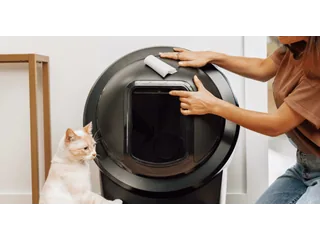 Una persona limpia con un paño el interior de un Litter-Robot 4 negro mientras un gato permanece junto a la entrada; fotografía oficial de Whisker.