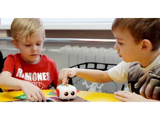 Dos niños programan un Tale-Bot Pro blanco con accesorios rojos sobre un tapete, en una fotografía oficial de MatataStudio.