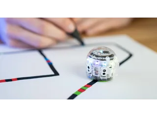 Fotografía oficial de un Ozobot Evo transparente sobre una línea con códigos de colores, mientras una mano dibuja el recorrido al fondo.