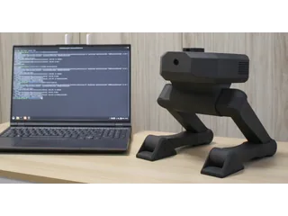Fotografía oficial del robot bípedo Bimo sentado sobre una mesa junto a un ordenador portátil.